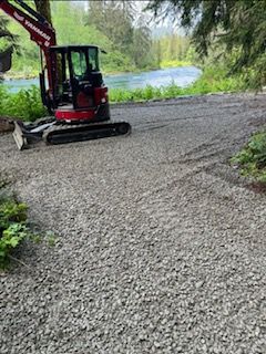 A red Yanmar excavator parked on a gravel area next to a river surrounded by trees.