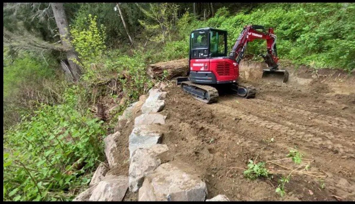A red excavator sits on a dirt path next to a newly constructed stone retaining wall on a hillside.