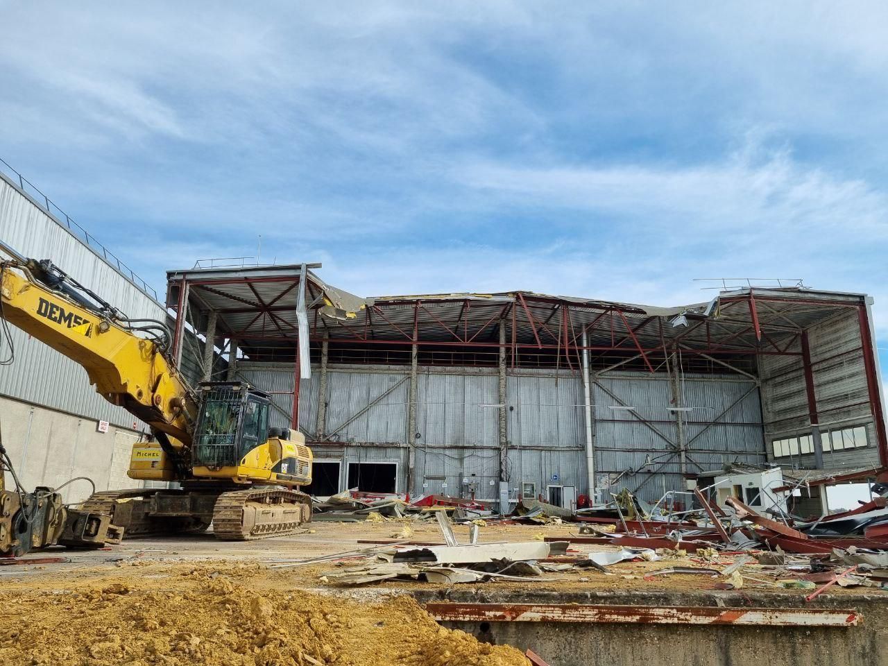 Un grand bâtiment est en cours de démolition avec une excavatrice jaune au premier plan.