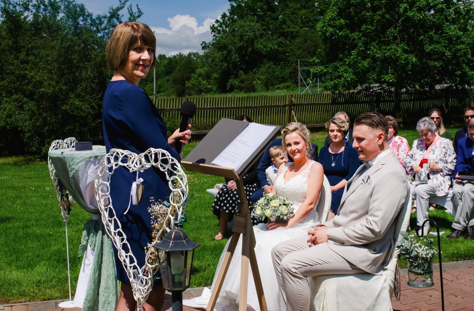 Eine Frau hält bei einer Hochzeitszeremonie vor Braut und Bräutigam eine Rede.