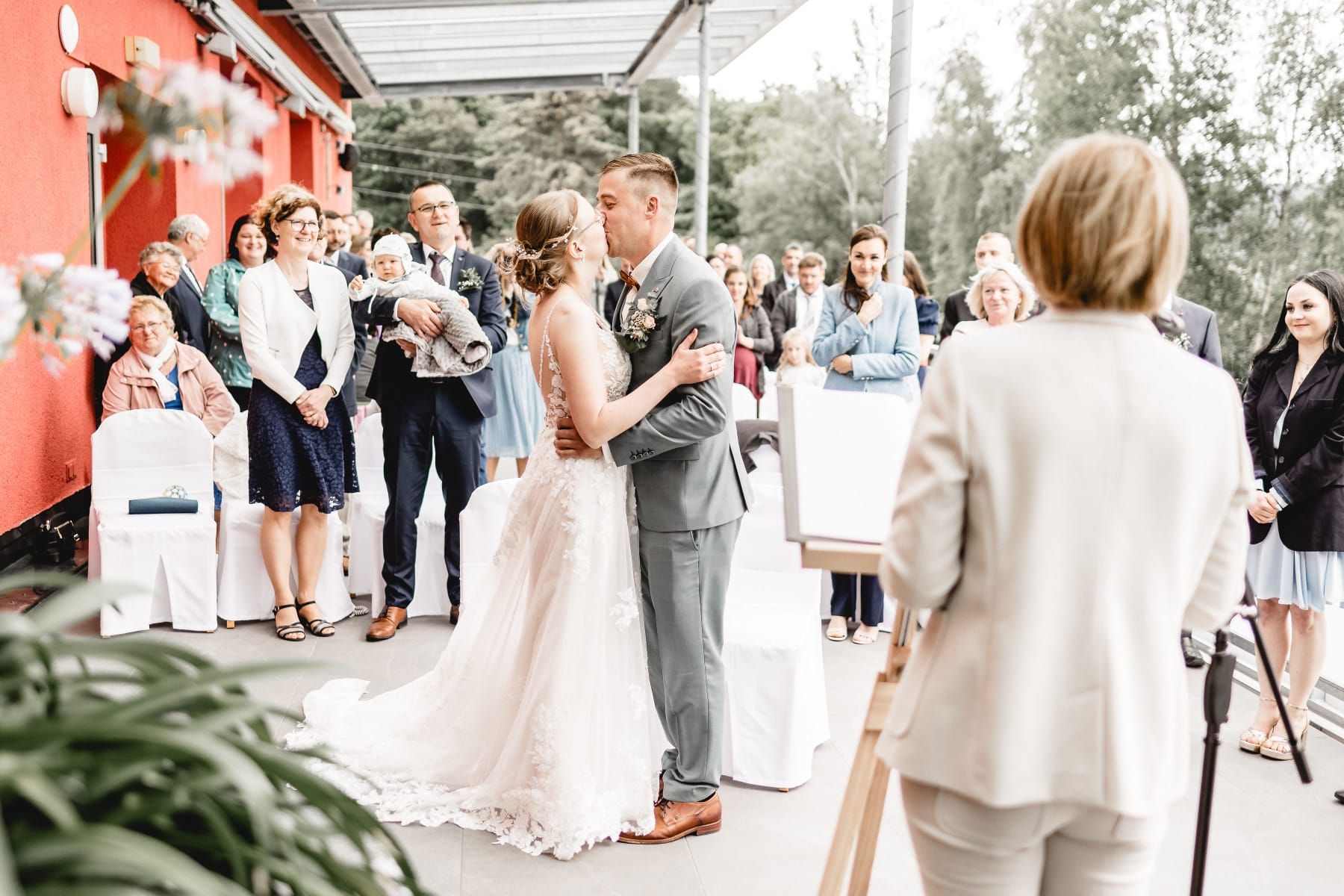 Eine Braut und ein Bräutigam küssen sich während ihrer Hochzeitszeremonie, während eine Frau hinter ihnen steht.