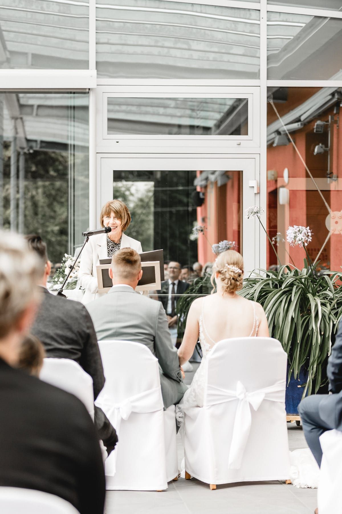 Bei einer Hochzeitszeremonie sitzen Braut und Bräutigam auf weißen Stühlen, während eine Frau in ein Mikrofon spricht.