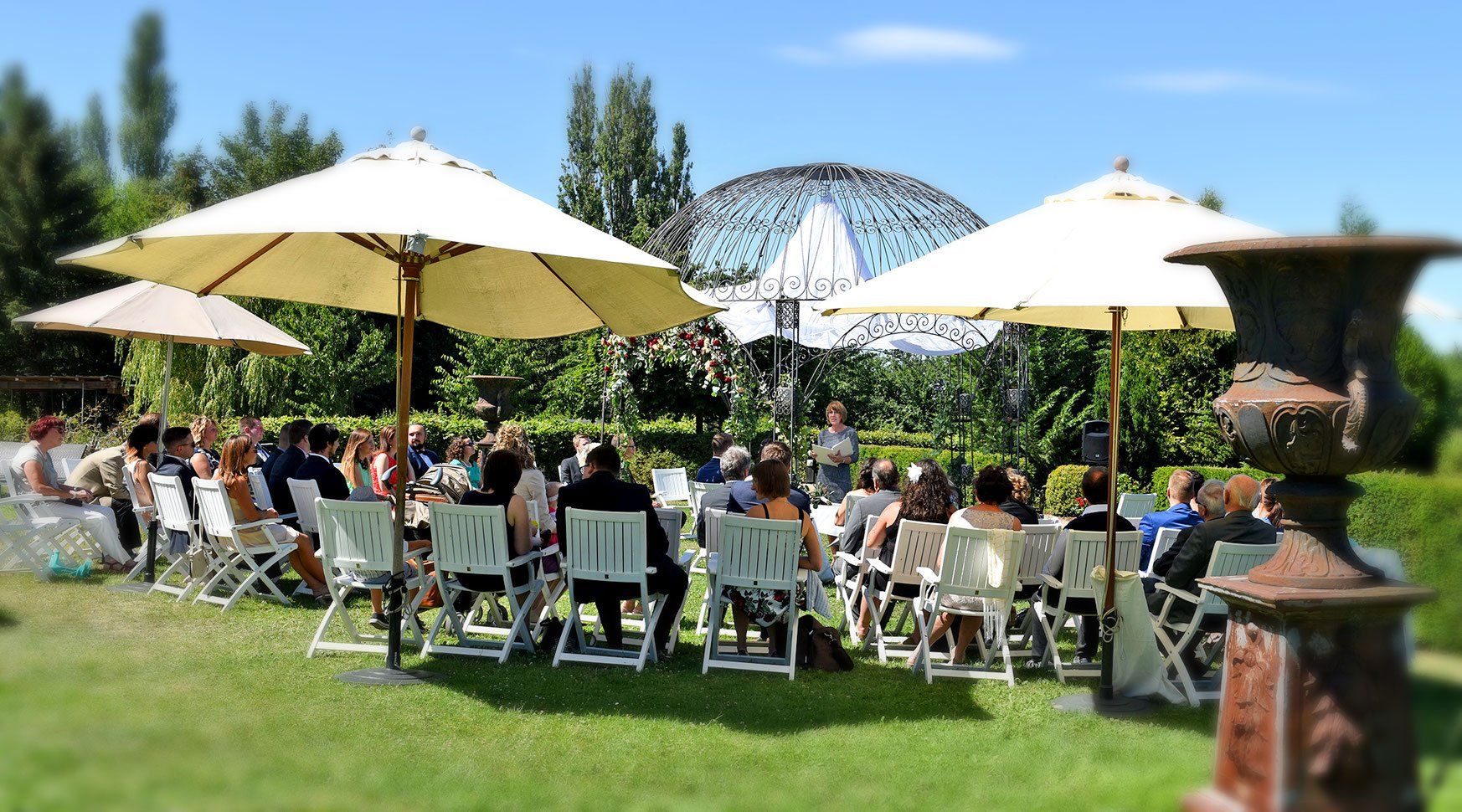 Eine Gruppe von Menschen sitzt während der Trauung unter Sonnenschirmen in einem Park