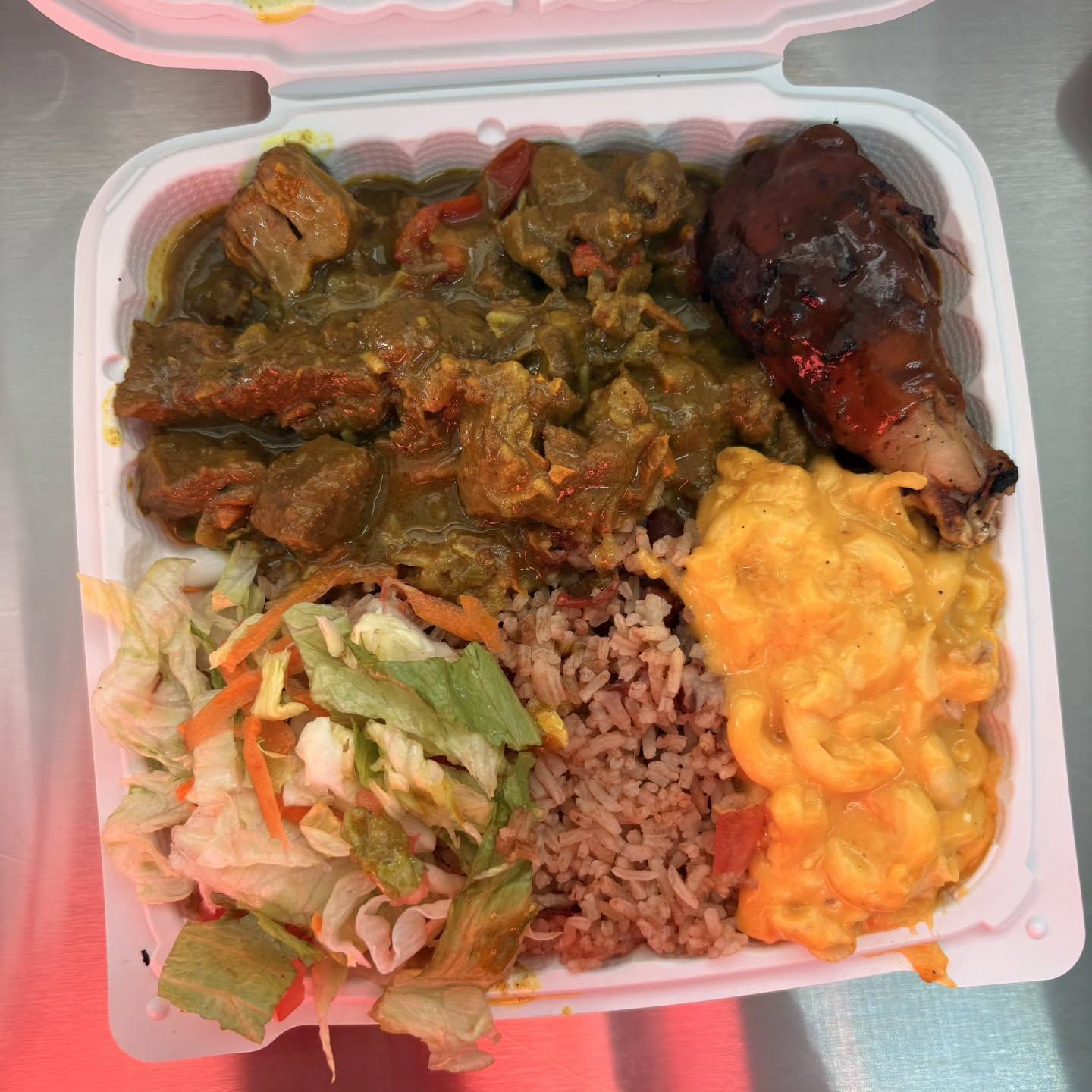 Takeout container with curry meat, jerk chicken, rice, macaroni and cheese, and salad.