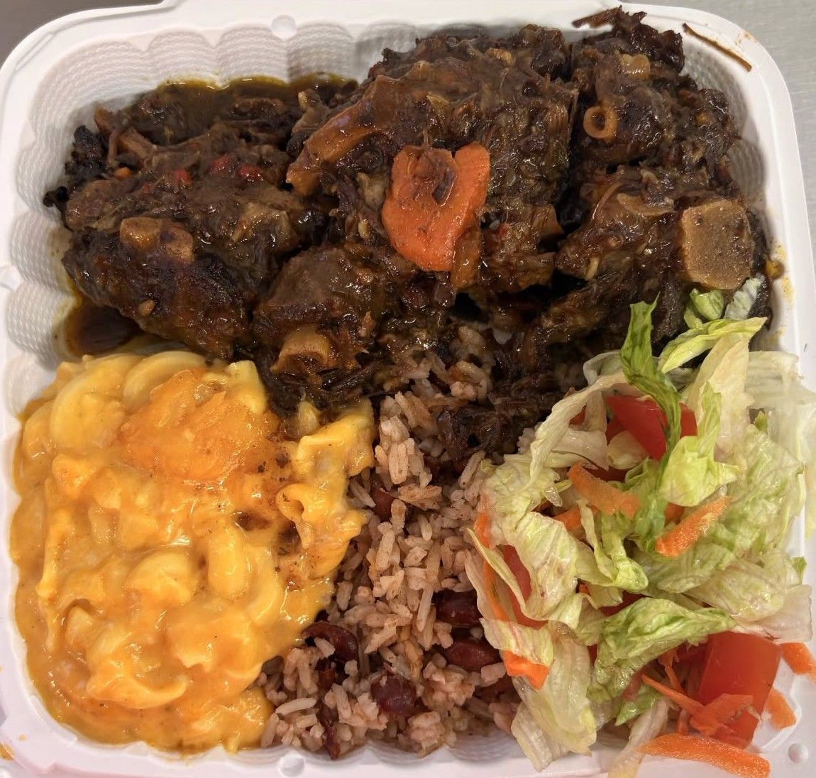Oxtail meal with mac and cheese, rice and peas, and salad in a styrofoam container.