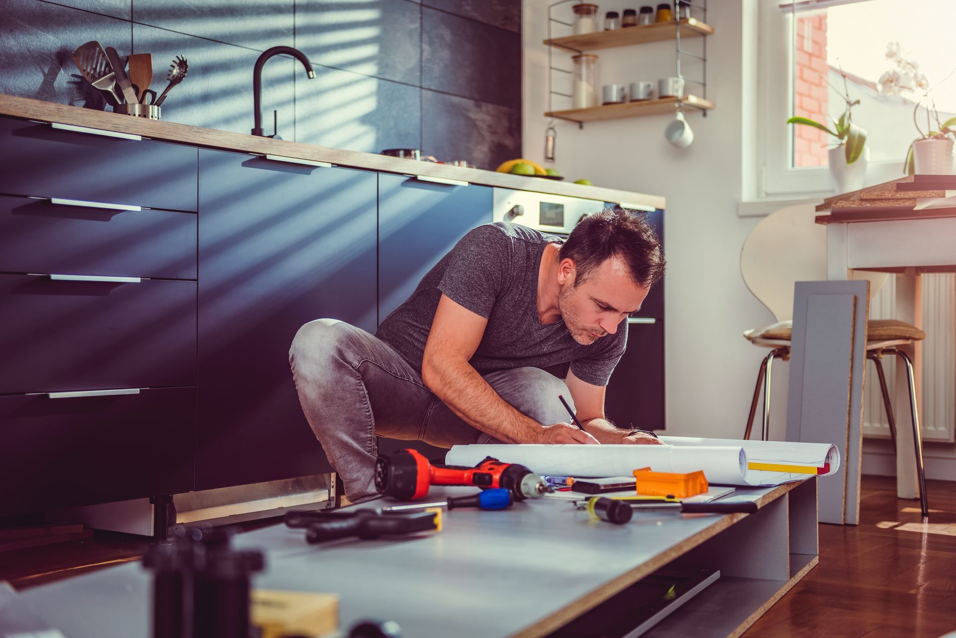 A man is kneeling down in a kitchen working on a piece of wood.