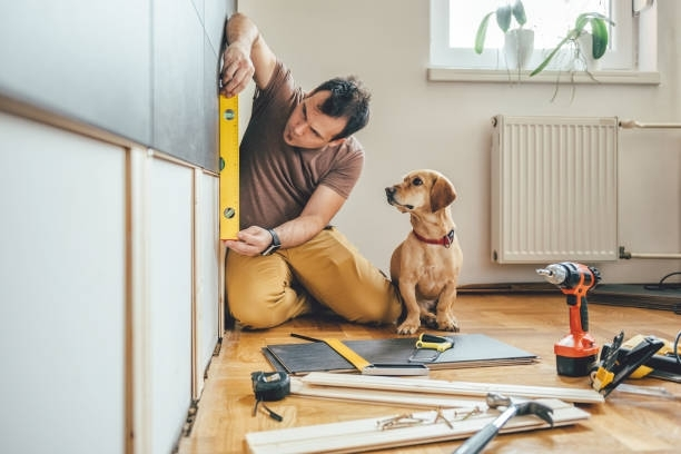A man is kneeling on the floor with a dog while measuring a wall.