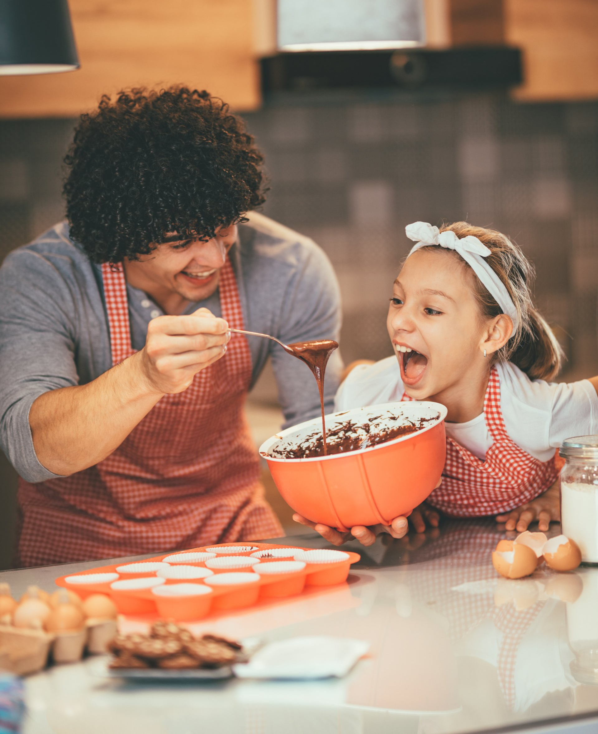 A father and daughter bonding while baking cupcakes. The dad is stirring the batter while the daughter looks on with excitement. Both are wearing aprons and have big smiles, showing the love and connection they share in this special moment.