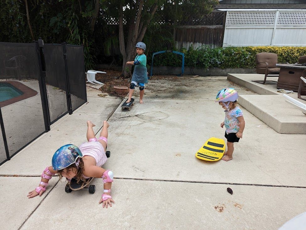 3 children of varying ages play on skateboards in their backyard