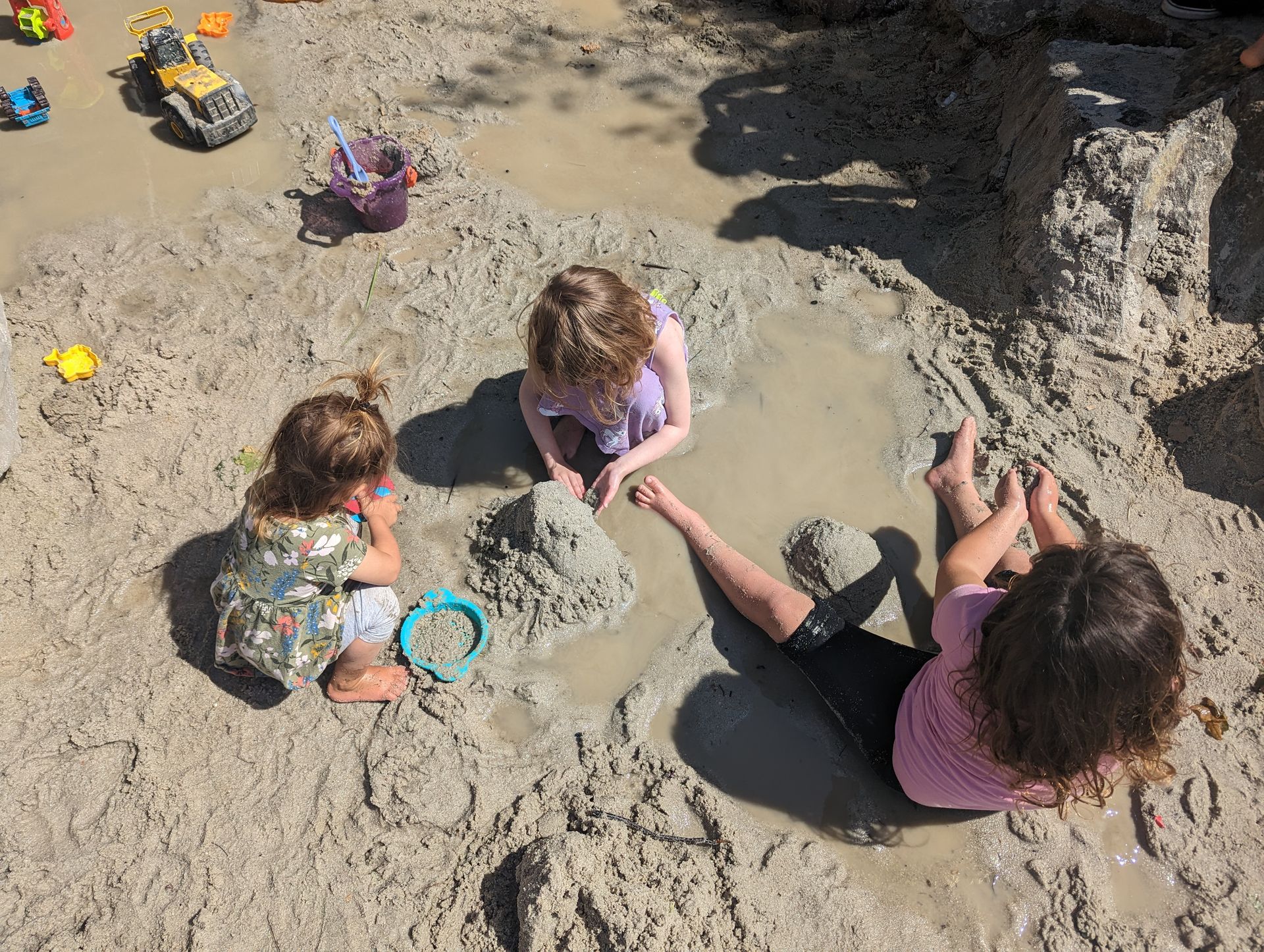 3 children sit in a mud pit building sandcastles