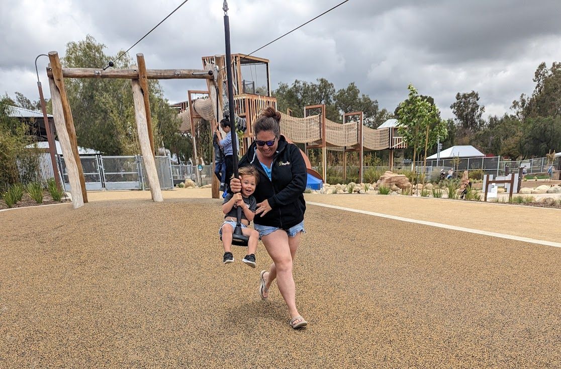 A mom runs beside her child riding a zipline at a park