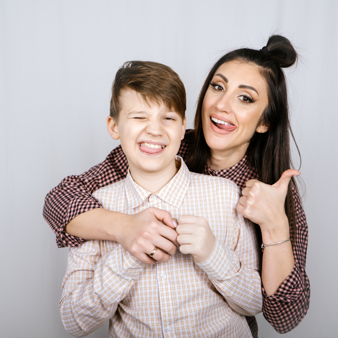 A mom and young teen child making goofy, happy faces in to the camera while embracing