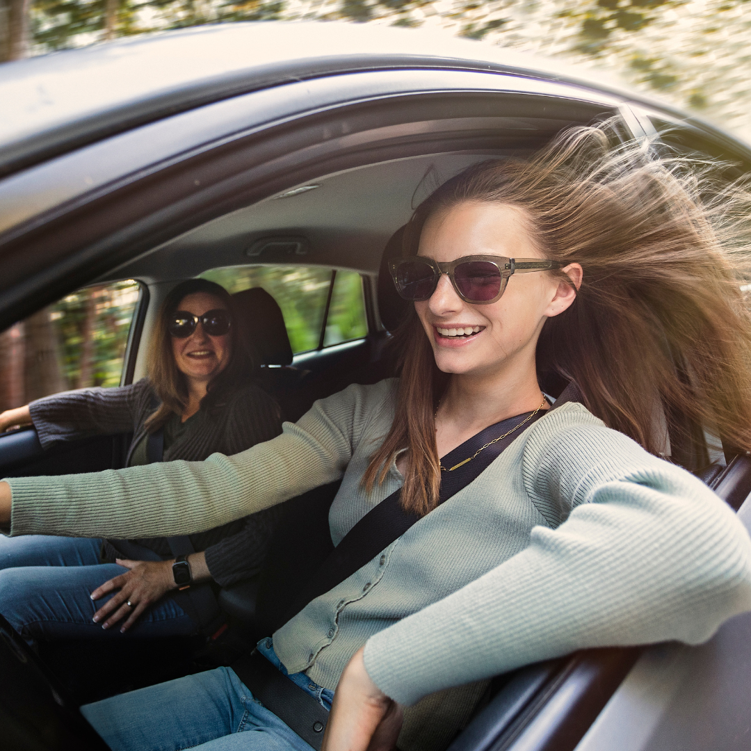 A teen girl driving with the window down with her mother looking at her adoringly from the passenger seat