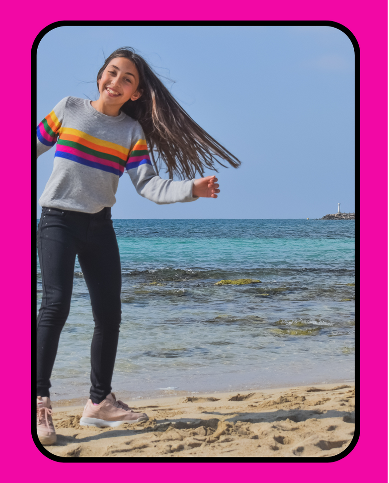 A young teen in a sweater and jeans standing smiling on a beach in front of the ocean