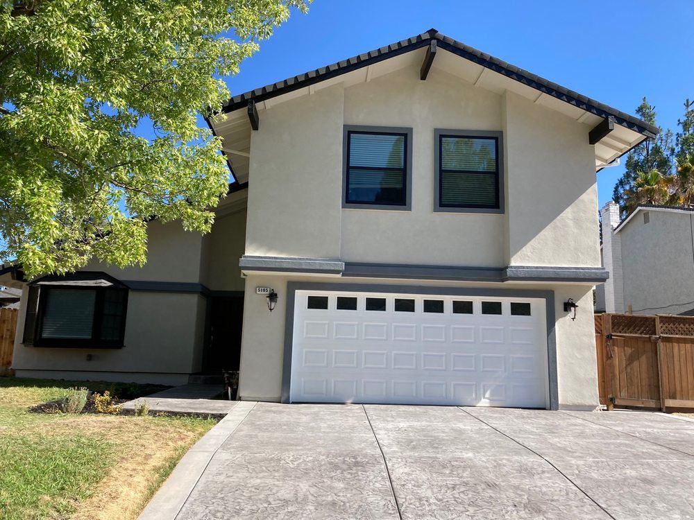 Suburban two-story house with white garage, light stucco exterior, and driveway under a blue sky