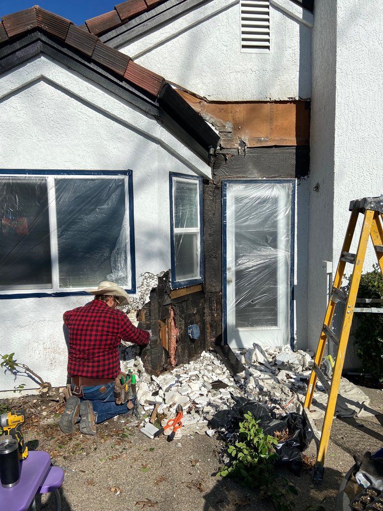 Worker in red plaid jacket repairing damaged exterior wall beside a ladder and debris pile