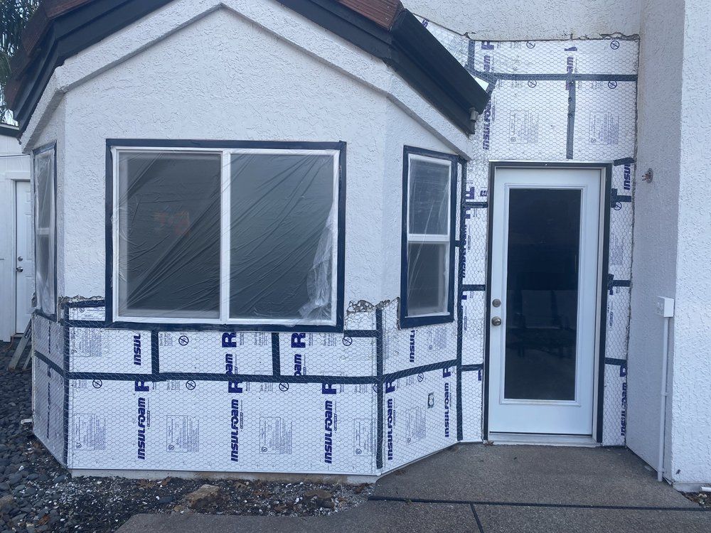 Exterior of a small house entrance with white siding, bay window, and a glass door under construction wrap