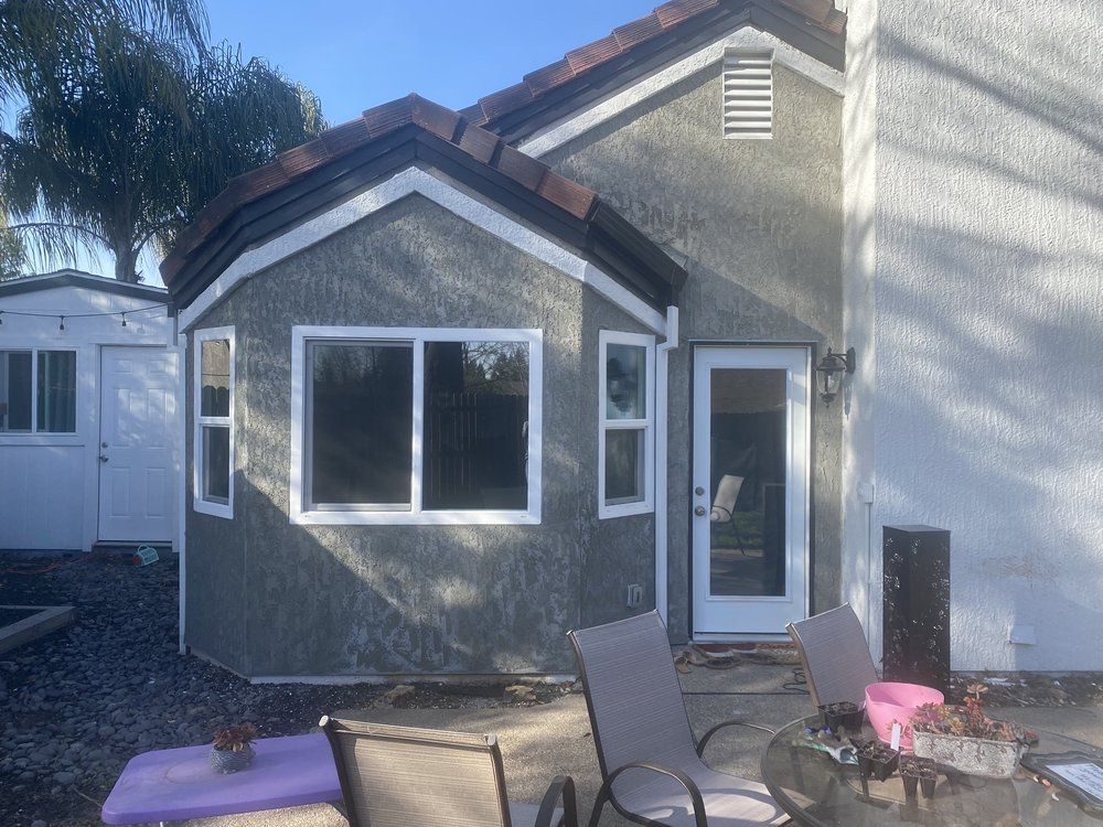 Backyard patio with a stucco house, large window, glass door, and outdoor chairs in sunlight