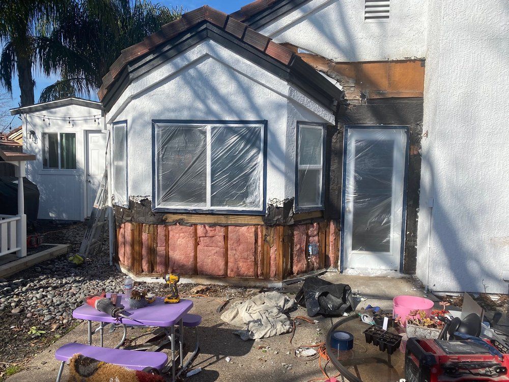 Backyard patio with a white house, a bay window, and outdoor tables, chairs, and tools scattered on the ground