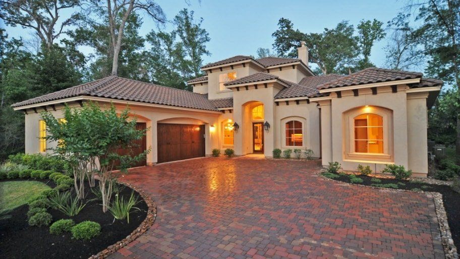 Luxury stucco house at dusk with warm-lit windows, tiled roof, and brick driveway, surrounded by trees