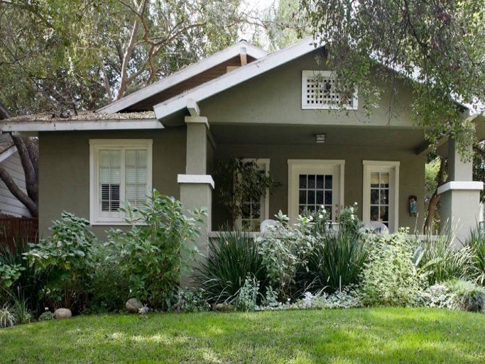 Small green house with white trim and a front porch, surrounded by lush garden plants.