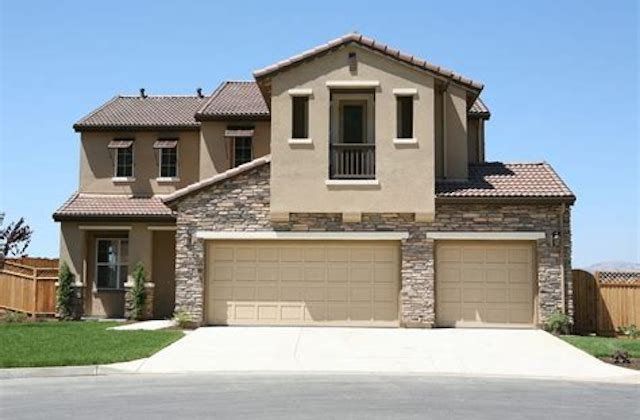 Large two-story suburban house with beige stucco, stone accents, and a three-car garage under a blue sky