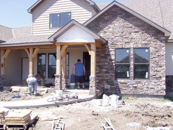 Workers building a stone-faced house exterior with porch framing and construction materials scattered outside