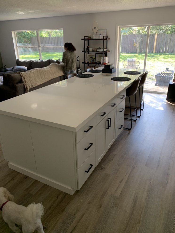 Bright kitchen with a large white island, stools, a person by the window, and a small white dog on the floor