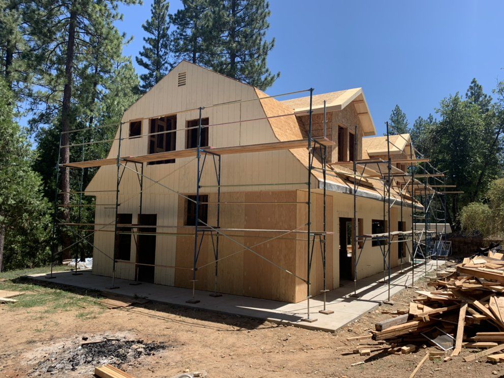 Wood-framed house under construction with scaffolding, set among tall pine trees under a clear blue sky.