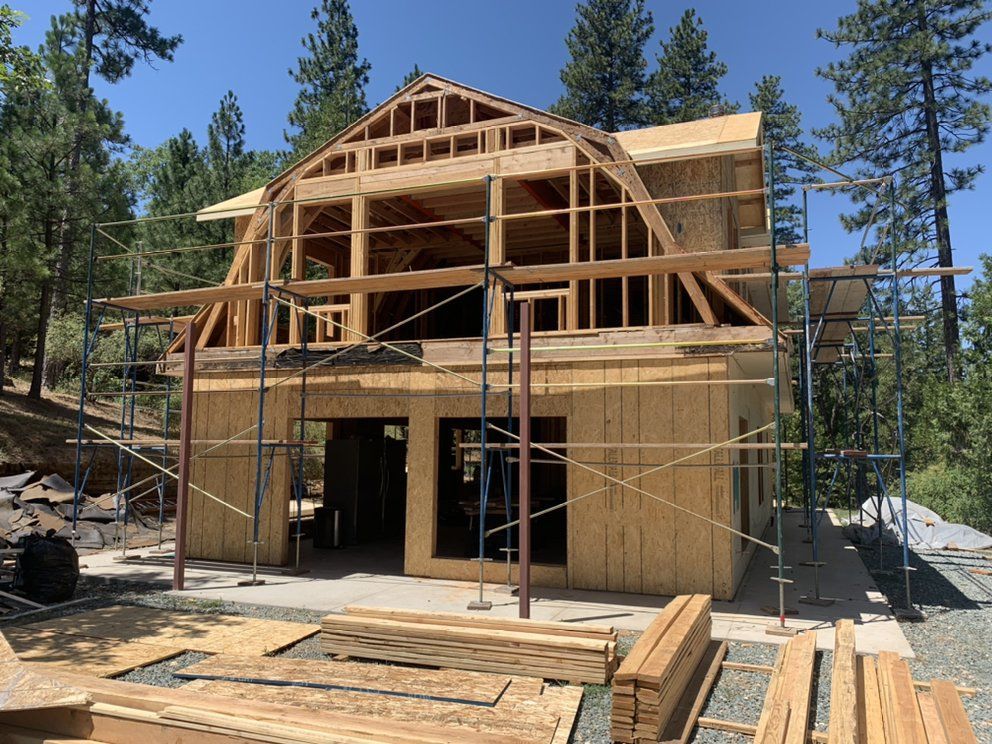 Two-story house under construction with wood framing and scaffolding in a wooded area