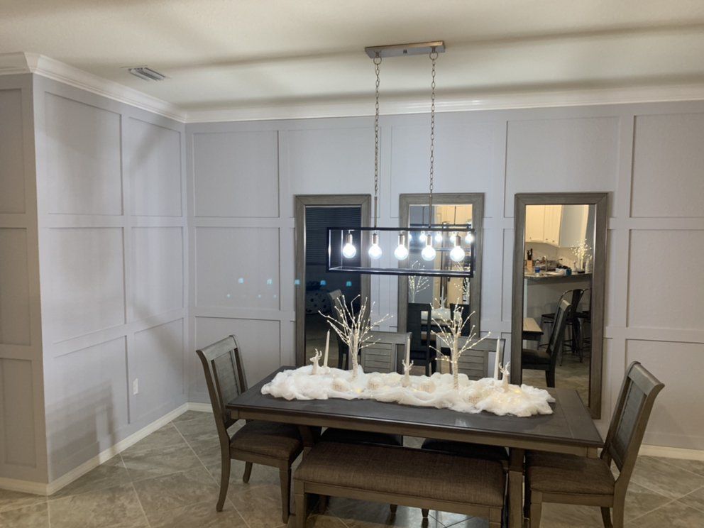 Modern dining room with a wooden table, chairs, and a chandelier over a white paneled wall.