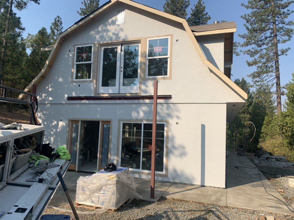 Two-story house under construction with white exterior, large windows, and scaffolding in a wooded area