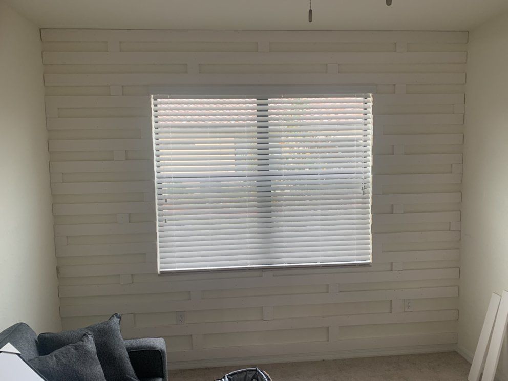 Empty living room with white paneled wall and a window with blinds, gray sofa in the corner.