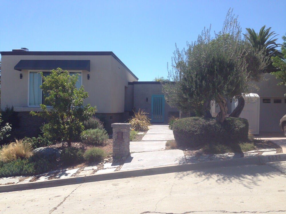 Single-story beige house with a front walkway, shrubs, and a tree under a clear blue sky