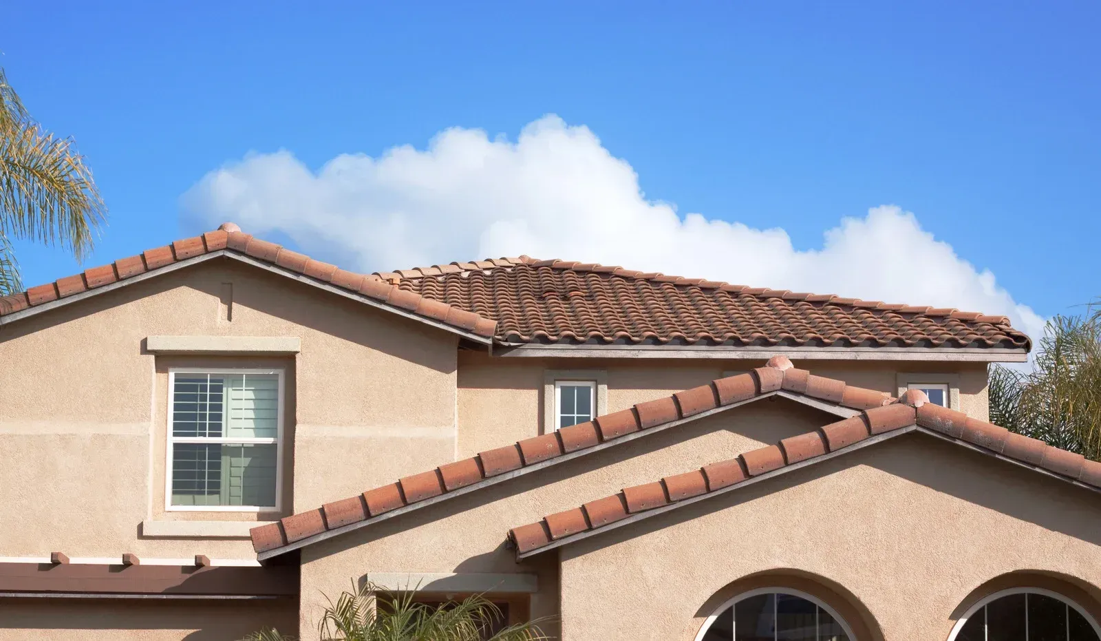 Terracotta-tiled house roof and beige stucco exterior under a blue sky with clouds