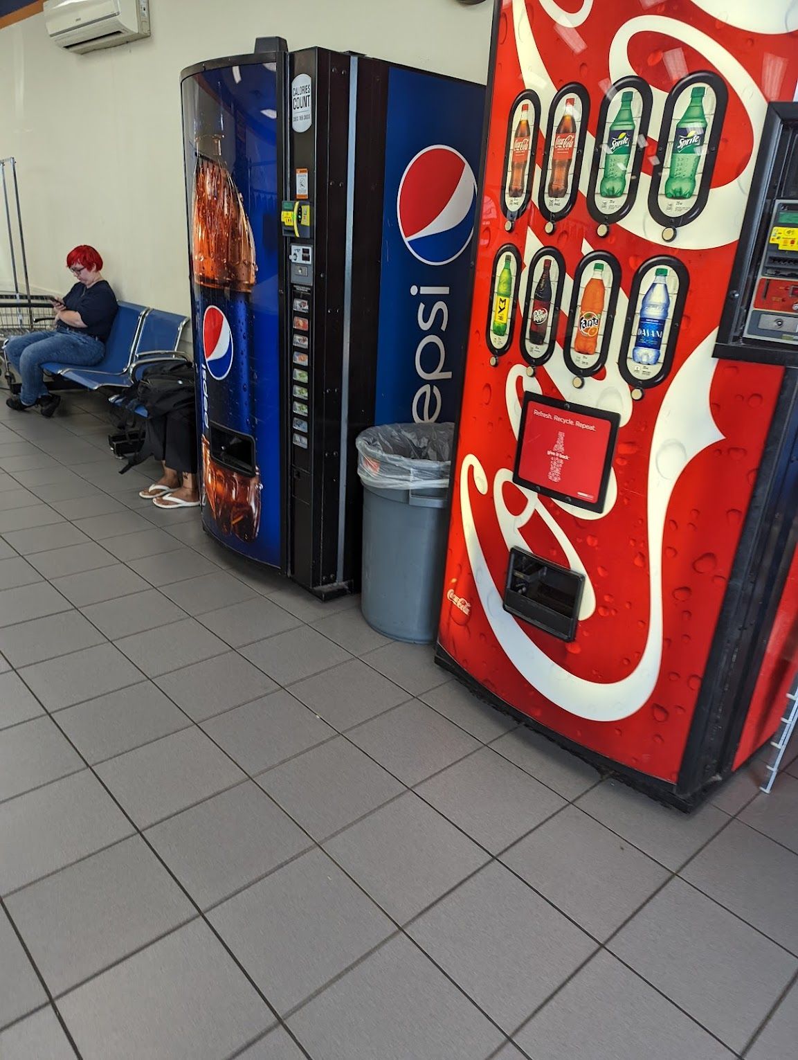 A woman sits on a bench next to a pepsi and coca cola vending machine