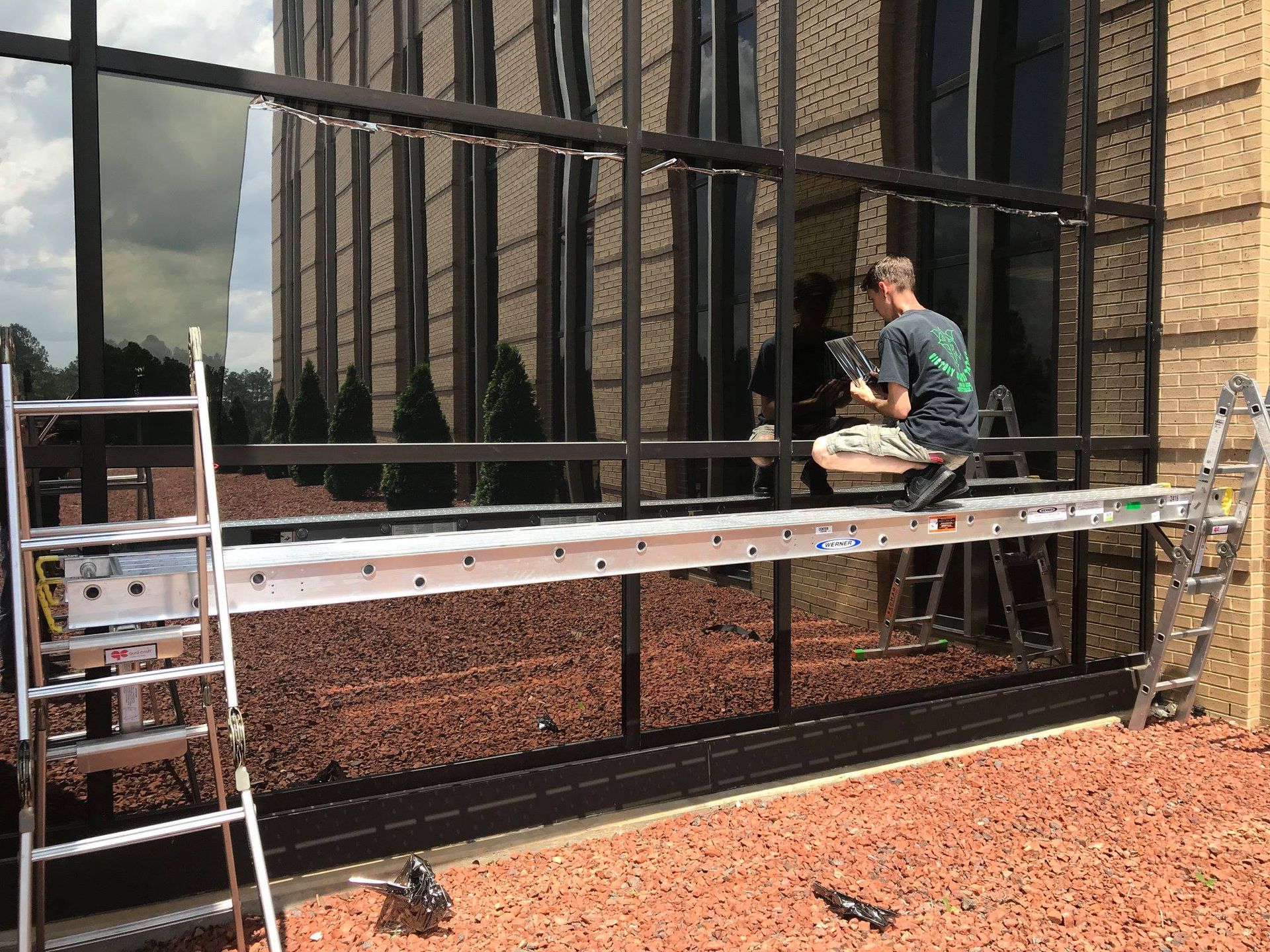 A worker sits on a metal scaffolding plank suspended between two ladders while installing glass on a commercial building.
