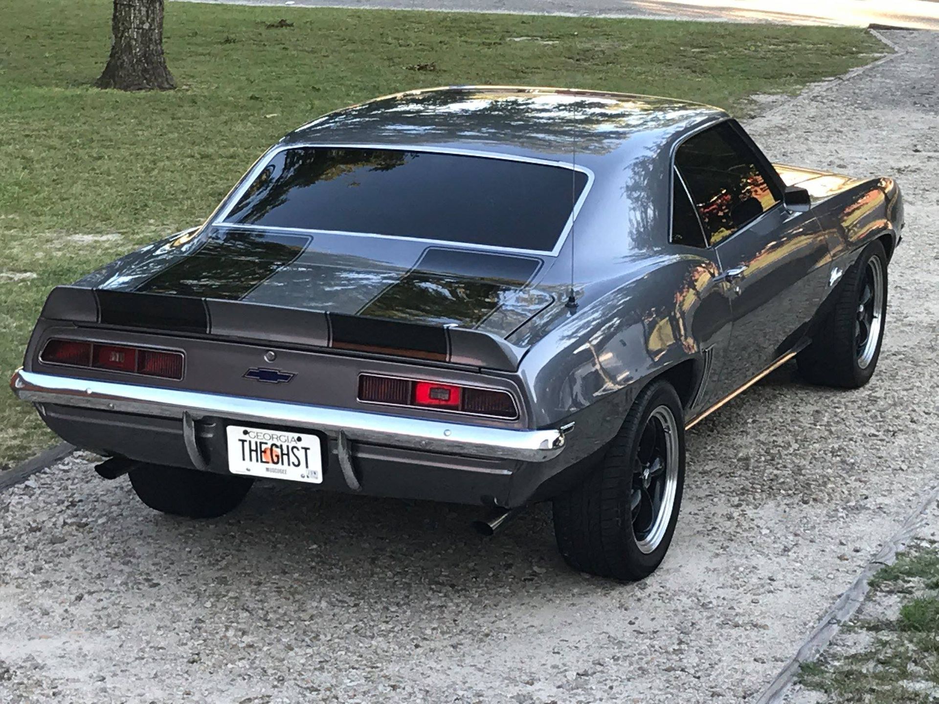 A grey vintage Chevrolet Camaro parked on a gravel driveway near a grassy area.