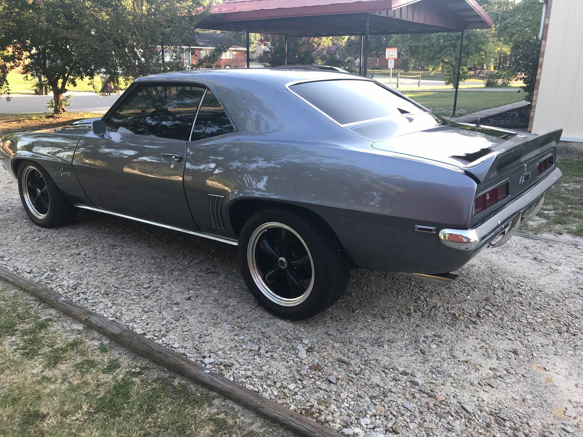 A gray vintage Chevrolet Camaro parked on a gravel driveway under a metal carport.