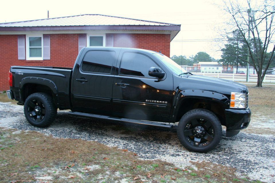 A black pickup truck with tinted windows and custom wheels parked on a gravel driveway in front of a red brick building.
