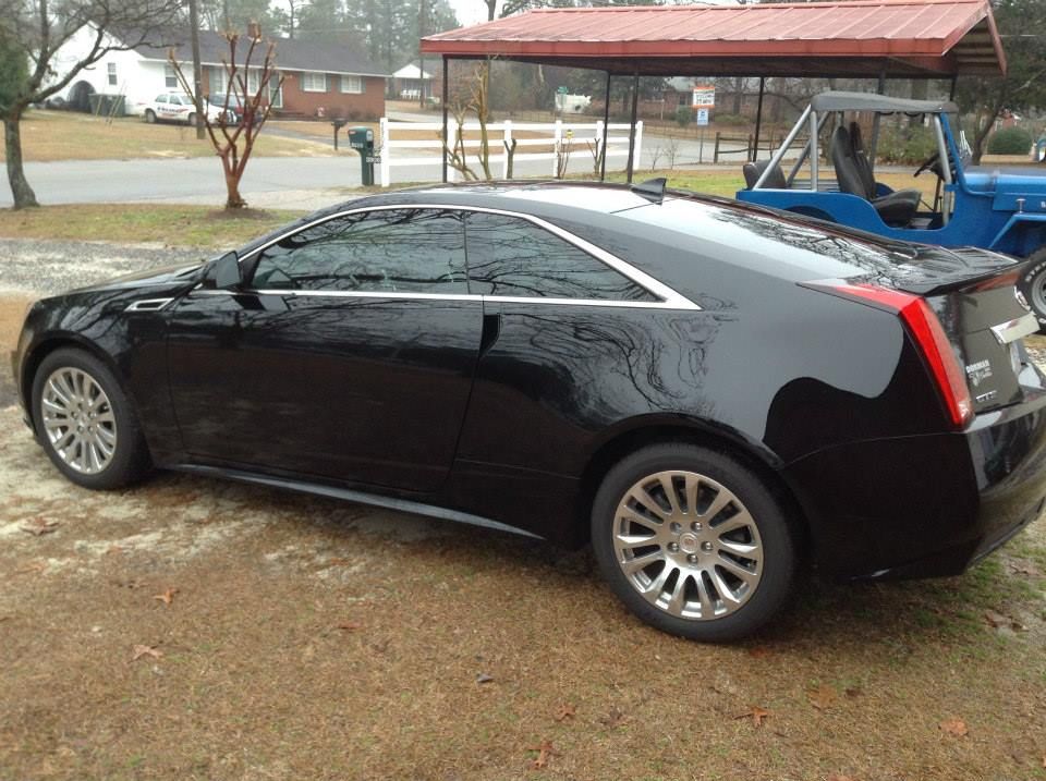 A black Cadillac CTS coupe parked on a grassy lot next to a carport and a blue utility vehicle.