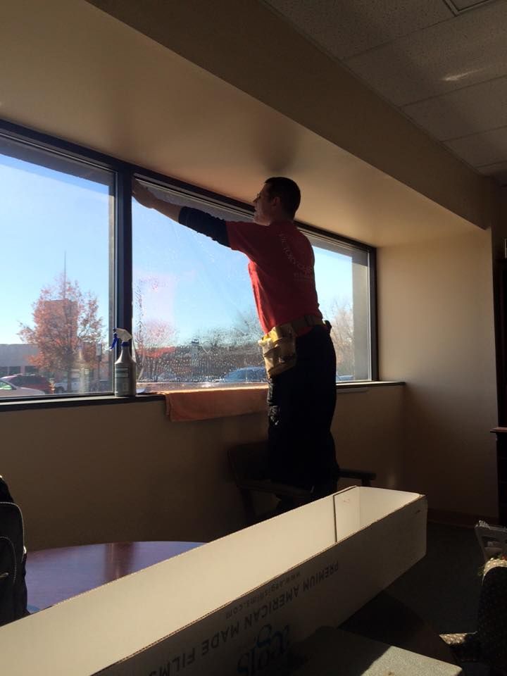 A person in a red shirt stands on a step stool while applying adhesive film to a large office window.
