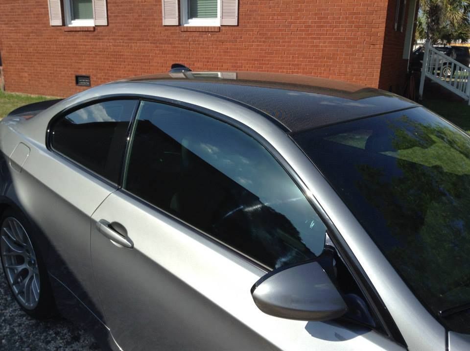 A silver two-door coupe parked in front of a red brick house features a dark, glossy roof and tinted windows.