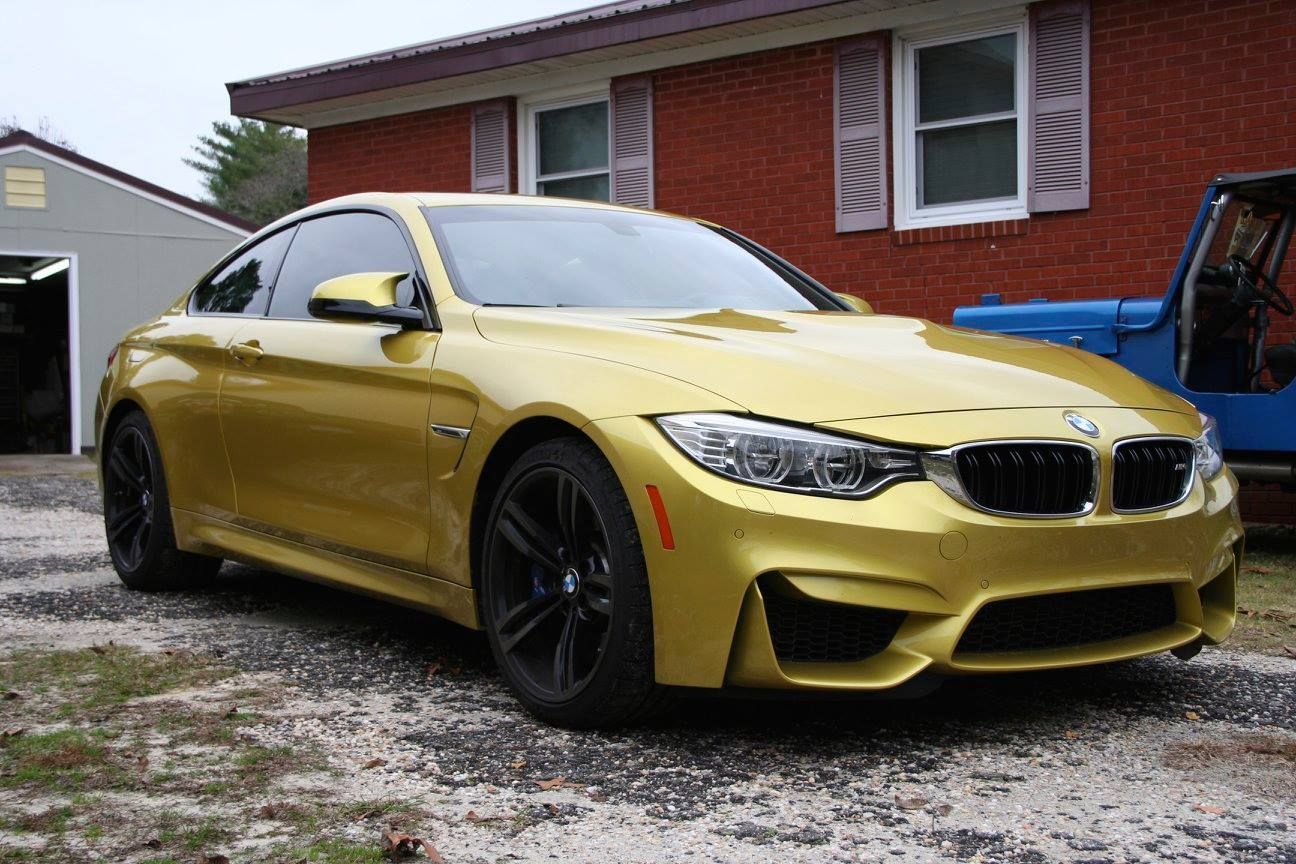 A yellow BMW M4 coupe parked on a gravel driveway in front of a red brick house.