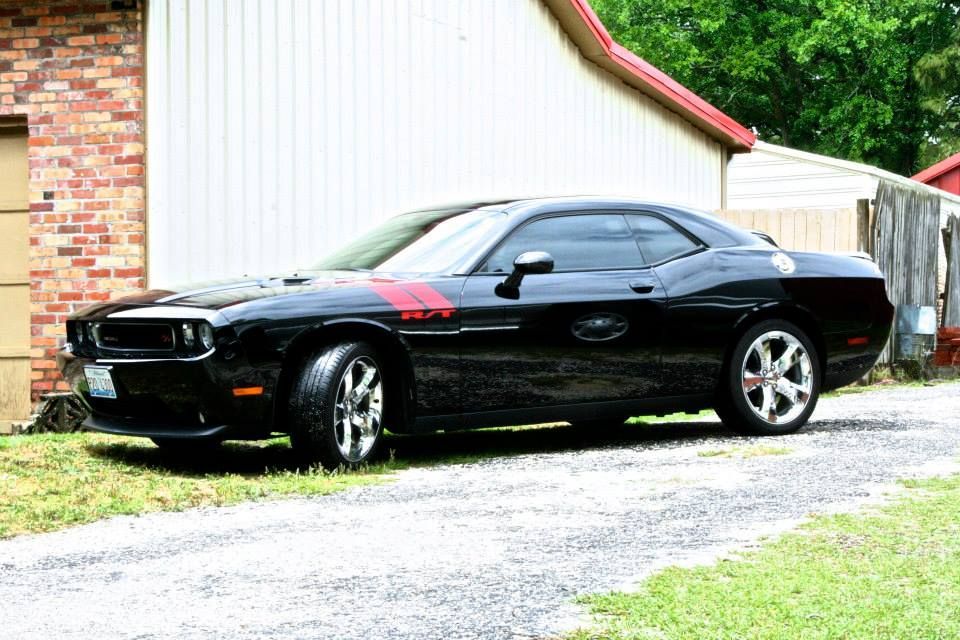 A black Dodge Challenger R/T parked on a gravel driveway in front of a brick building.