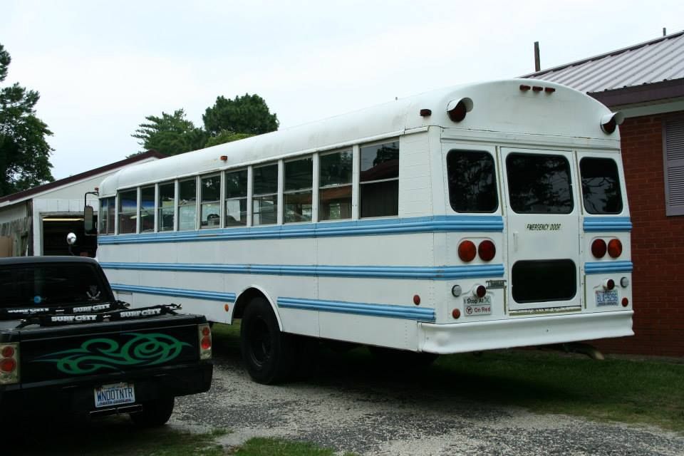 A white bus with blue horizontal stripes parked next to a black pickup truck on a gravel driveway.