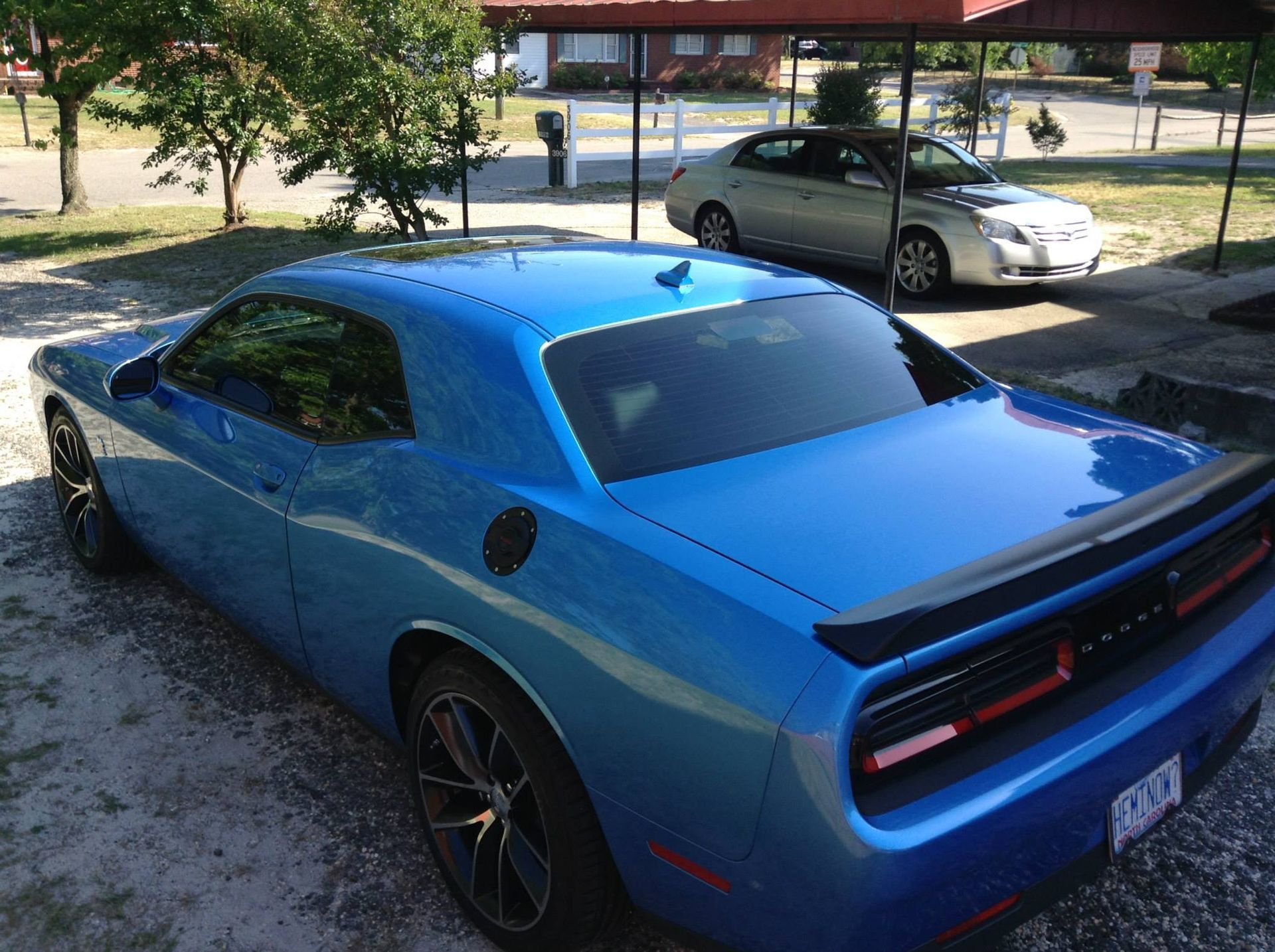 A bright blue Dodge Challenger parked outdoors under a metal carport, with a silver sedan parked behind it.