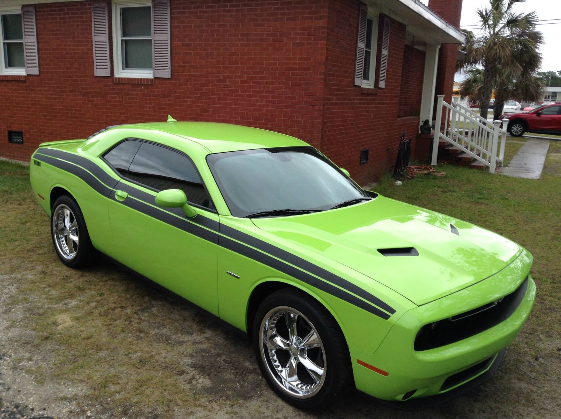 A bright lime-green Dodge Challenger with black side-stripes parked on grass in front of a red brick house.