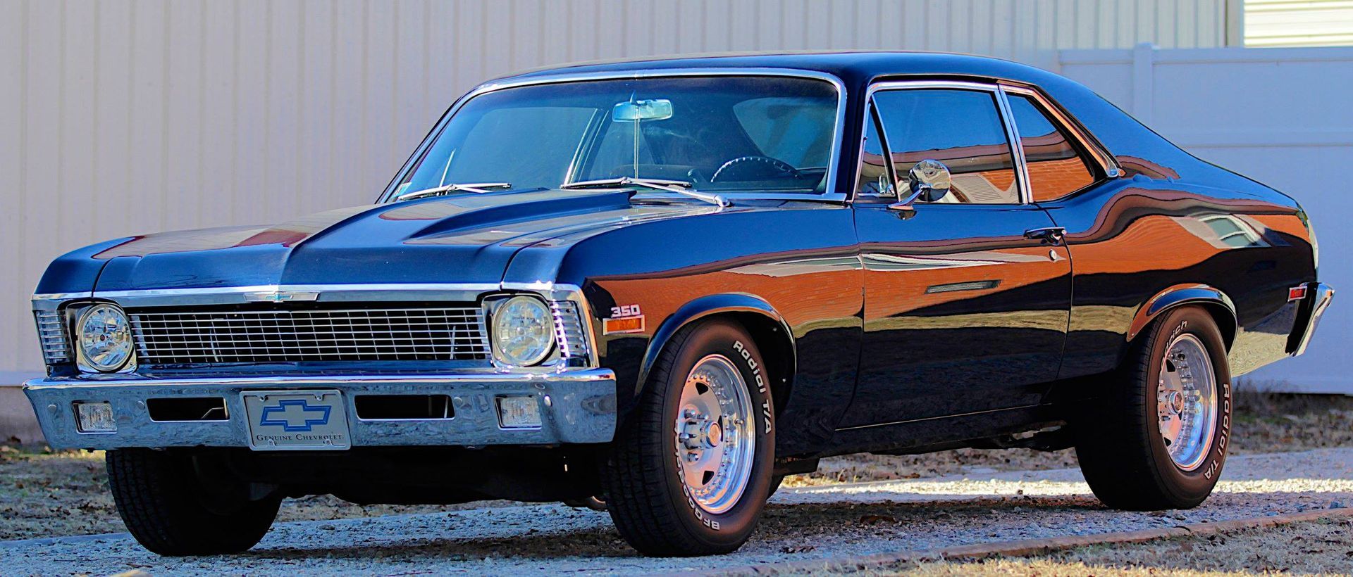 A dark blue, restored Chevrolet Nova muscle car parked on a gravel driveway in front of a white wall.