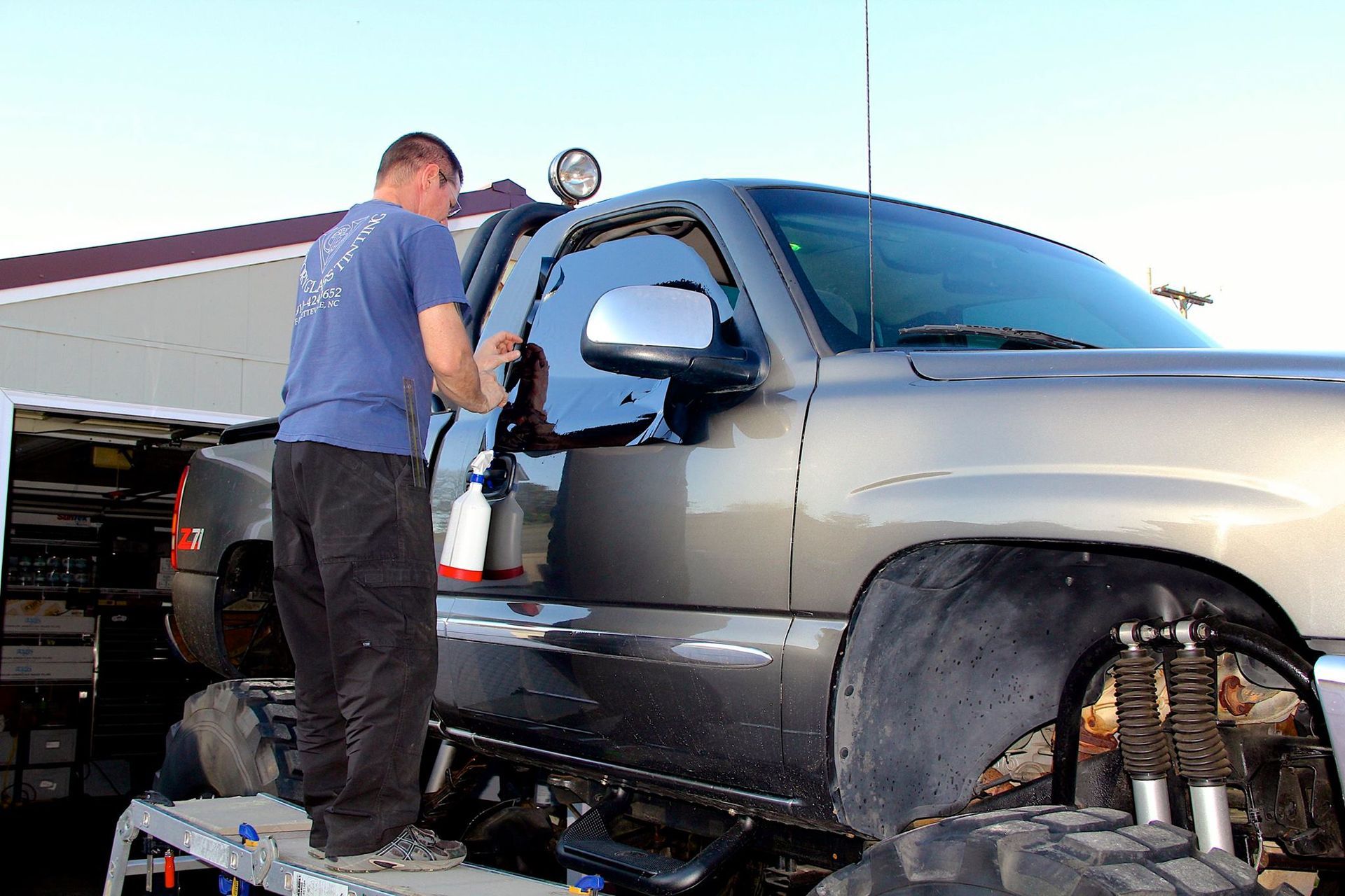 A person stands on a small step ladder, cleaning the window of a large, lifted gray truck outdoors.